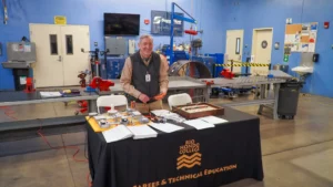 A smiling person stands behind a table with Rio Hondo College Career & Technical Education materials in a workshop filled with various tools and equipment.