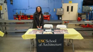 A person stands behind a table covered with brochures and a USC School of Architecture banner in a room with workshop equipment in the background.