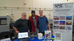 Three men stand together at a booth displaying engineering services, with a laptop and various engineering models on the table. A banner behind them reads "SPEC Services: Planning and Engineering for Essential Energy Systems.
