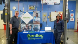 Two individuals stand behind a Bentley table display at an event. The table features promotional materials and a laptop, with posters in the background. The setting is a room with machinery and safety equipment.