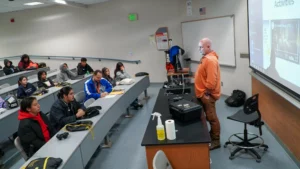 A teacher in an orange hoodie stands in front of a classroom, speaking to students seated at desks. A projector displays a presentation on the wall. Classroom supplies are on the desk.