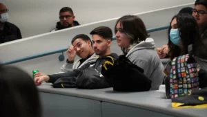 A group of students sits in a classroom with some wearing masks and casual clothing, while one student appears to wipe their eye.