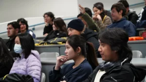 A group of students attentively listen in a classroom, some taking notes and one student raising their hand. Several students are wearing jackets, and one student in the foreground is wearing a face mask.