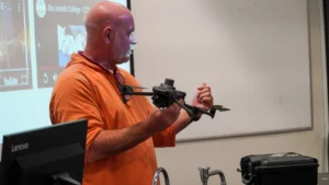 A person in an orange shirt holds a drone and gestures with their right hand during a presentation in front of a projector screen.