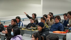 A group of students sit in a lecture hall, listening attentively. One person raises their hand, while others look engaged in the session. Some students wear jackets and one wears a face mask.