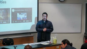 A person standing in front of a projected screen giving a presentation in a classroom. There's a whiteboard, a table with papers, cleaning supplies, and students seated.