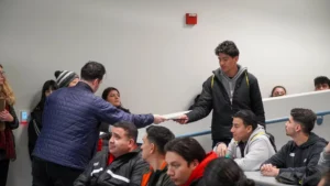 A man in a blue jacket hands a certificate to a young man in a grey hoodie in a classroom with several seated students.
