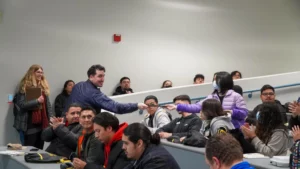 A man in a blue jacket hands a piece of paper to a woman in a purple jacket in a lecture hall. Several people are seated, watching and clapping.