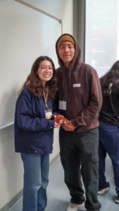 Two people standing next to each other in a casual setting. The person on the left is smiling, wearing a blue jacket, and holding a small item. The person on the right is wearing a brown hoodie and a yellow beanie.