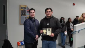 Two men are standing indoors. The man on the right is holding a plaque with a gold award. Several people are standing in the background.
