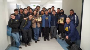 A group of young people and two adults pose in a hallway. Some hold plaques and trophies.