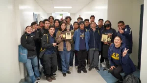 A group of people stand in a hallway, smiling and holding awards. Some are wearing matching jackets and several are giving peace signs.