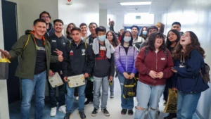 A group of diverse students stands in a hallway, some holding certificates and bags. They appear cheerful, with a few wearing masks and several smiling or laughing.