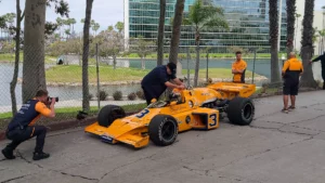 Pit crew in orange uniforms working on a yellow number 3 race car near a palm-lined waterway, with one member photographing the scene.