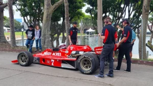 A red vintage racing car with "trust acres" and "good year" logos on display, surrounded by four men in casual attire and headsets, in a park with a fence and trees.