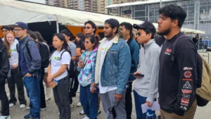 A group of diverse young adults standing in line at an outdoor event, looking ahead attentively.