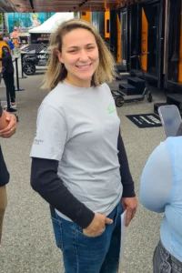 A smiling woman in a white long-sleeve shirt and denim jeans standing in an outdoor setting with vehicles and people in the background.