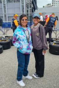 Two women smiling at the camera, one in a tie-dye hoodie and the other in a brown sweatshirt, standing by a stack of racing tires at a sports event.