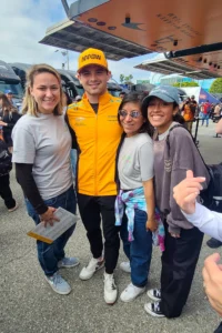 Three fans posing with a professional race car driver wearing a team uniform and cap, all smiling, at a racing event.