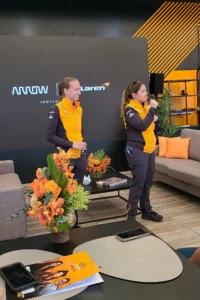 Two women in mclaren racing team uniforms speaking at an event, with one woman addressing an audience while the other listens, standing near a table with floral arrangements.