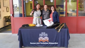 Three smiling women stand behind a table with a "Rio Hondo College, Career and Technical Education" tablecloth. The table has various items on it, including cords and bags.
