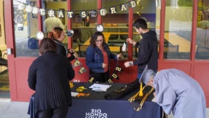 People arranging decorations and items on a table covered with a "Rio Hondo College" cloth, under a banner that says "Congrats Grad.