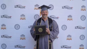 A man in a graduation cap and gown smiling and holding a diploma in front of a backdrop with "Automotive Technology" and "Rio Hondo College" logos.