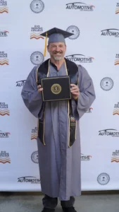 A man in a graduation gown and cap holds a diploma and smiles at the camera, standing in front of a backdrop featuring logos from Rio Hondo College and automotive technology.