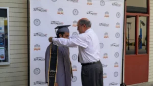 A graduate in a gray cap and gown stands as an older man adjusts his honor cord in front of a step-and-repeat banner at an automotive college event.