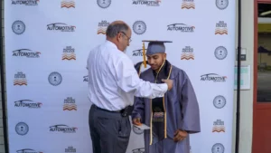 A man in a white shirt adjusts the graduation stole of a student wearing a cap and gown. A backdrop with "Automotive Technology" logos is in the background.