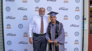A man in a white shirt with a colorful tie stands next to a young graduate in a cap and gown. They pose for a photo in front of a backdrop with various logos, including "Automotive Technology" and "Rio Hondo College.