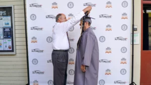 A man in a white shirt places a graduation stole around the neck of another man in a gray graduation gown and cap, standing in front of a backdrop with "Automotive" logos.