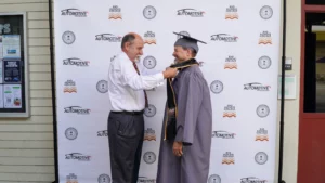 An older man in graduation robes receives a medal from another man in front of a backdrop with automotive and educational logos.