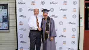 A graduate in a cap and gown shakes hands with an older man in a tie in front of a backdrop with logos, presumably at a graduation ceremony.