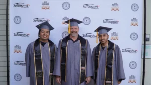 Three graduates wearing gray caps and gowns stand in front of a backdrop with "Automotive Technology" and "Rio Hondo College" logos.
