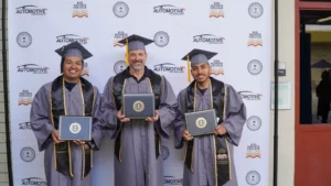 Three graduates in caps and gowns stand in front of an "Automotive Technology" backdrop, proudly holding their diplomas and smiling.