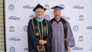 Two men in graduation gowns and caps stand in front of a backdrop with "Automotive Technology" and "Rio Hondo College" logos. One gown features "CSULA." Both men are smiling and holding diplomas.