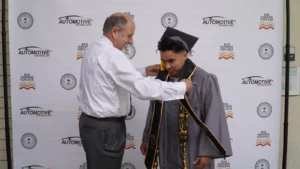 An older man adjusts the stole of a young man in a graduation cap and gown in front of an automotive technology and Rio Hondo College banner.