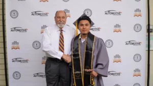 Two men stand side-by-side; one in a white shirt shaking hands with the other in a gray graduation gown and cap. They are in front of a backdrop with multiple logos including "Automotive Technology" and "Rio Hondo College.