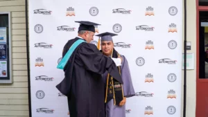 A graduate in a gray cap and gown stands next to an individual in academic regalia. They are in front of a backdrop with logos for an automotive technology program and a college.