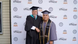 Two graduates in caps and gowns shake hands in front of a backdrop with "Automotive Technology" and "Rio Hondo College" logos.