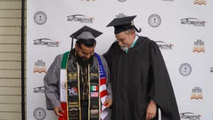 Graduate in cap and gown with decorative stoles stands next to a faculty member in regalia, both smiling and looking down, in front of a backdrop with "Automotive Technology" and "Rio Hondo College" logos.