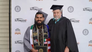 A graduate in cap and gown poses with a faculty member on a stage with an "Automotive Technology" background. The graduate holds a diploma and wears a sash adorned with flags and patches.