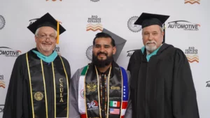 A graduate stands between two men in academic regalia, posing in front of a Rio Hondo College Automotive Technology backdrop. The graduate wears a decorated sash with various symbols and flags.