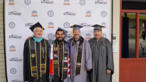 Four men in graduation gowns and caps stand in front of a backdrop with automotive-themed logos, smiling for a group photo. One man wears multiple sashes and stoles.