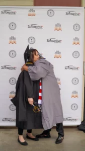 Two individuals in graduation gowns embrace in front of a backdrop with logos. One person holds a diploma and the other wears a scarf with red and white stripes.