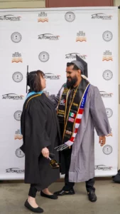 Two graduates in caps and gowns stand in front of a backdrop with "Automotive Technology" logos, facing and congratulating each other. The man wears a sash with flags and symbols.