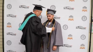 A graduate in a grey cap and gown receives a cord from an older man in a black robe and cap at a ceremony, with a backdrop featuring "Automotive Technology" and "Rio Hondo College" logos.