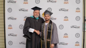 Two men in graduation caps and gowns stand in front of an "Automotive Technology" backdrop. The older man shakes hands with the younger man, who holds his diploma folder.