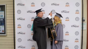A man in graduation attire adjusts the tassel on the cap of a woman also in graduation attire in front of a step-and-repeat banner featuring automotive and college logos.
